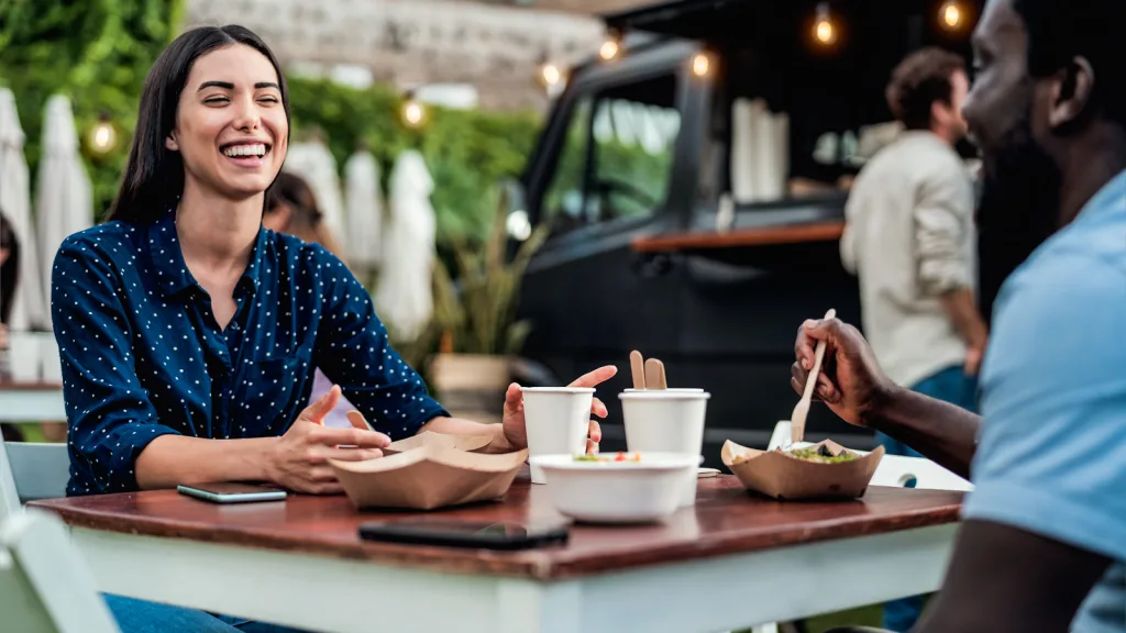 Florida-Woman in blue and white polka dot shirt and man in light blue shirt eating under string lights just before dawn at a food truck. They are both laughing and have their meal in various containers in front of them. New pet-friendly apartment homes for rent with private fenced backyards in Florida with pool, pickleball, dog park, gym, clubhouse and gated community. *No breed or weight restrictions. Yardly